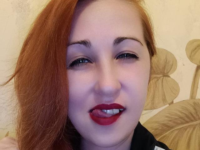 JillSilvies has red hair, light skin, and is sticking out her tongue with red lipstick and black eyeliner. Her eyebrows are slightly raised. She's wearing a black shirt.
