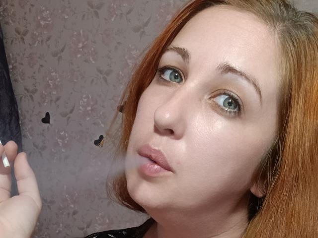 JillSilvies has pale skin, red hair, and green eyes with a slight pout, blowing a small cloud of breath near her pink lips. Her right hand is visible.