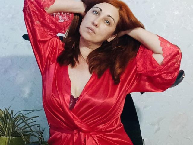 JillSilvies, con i capelli rossi, indossa una veste rossa lucida in pizzo, con le braccia alzate, le mani tra i capelli, le labbra leggermente aperte, che mostra il seno, seduta su una sedia nera..