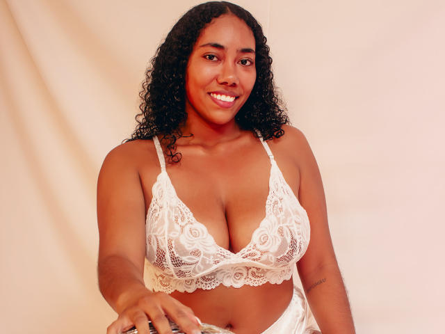 AishaRhodes with curly black hair, brown skin, and medium tits, wearing white lace bra and panties, kneeling with a large silver mirror ball, smiling.