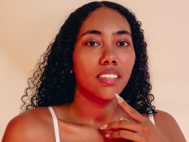 AishaRhodes with curly black hair, brown skin, white tank top, and pink lipstick, touching her chin with a finger, showing a small white dot on her lip.