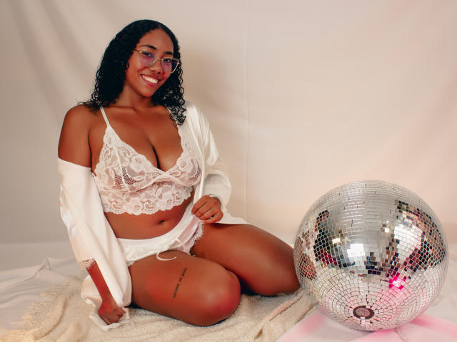 AishaRhodes with curly black hair, glasses, and red lipstick, wearing white lace bra and shorts, sitting on a towel, next to a silver mirror ball, smiling.