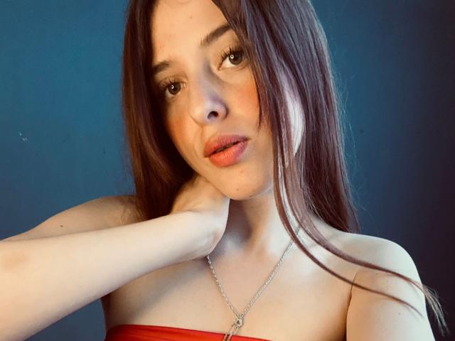 BlueViolette with long brown hair, fair skin, and pink lips, wearing a red strapless top, touching her neck, looking at the camera, wearing a silver necklace with a pendant.