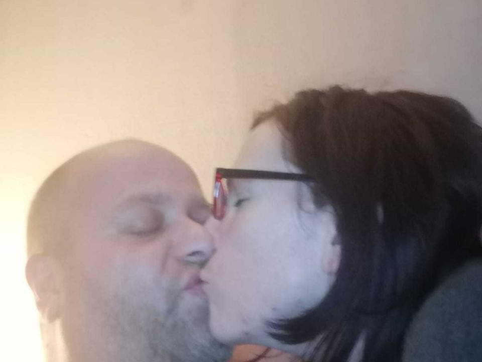 LoveJouse, met een kaal hoofd en baard, kust een vrouw met donker haar en een bril. Haar neus zit iets voor op de zijne. Haar lippen zijn op de zijne..