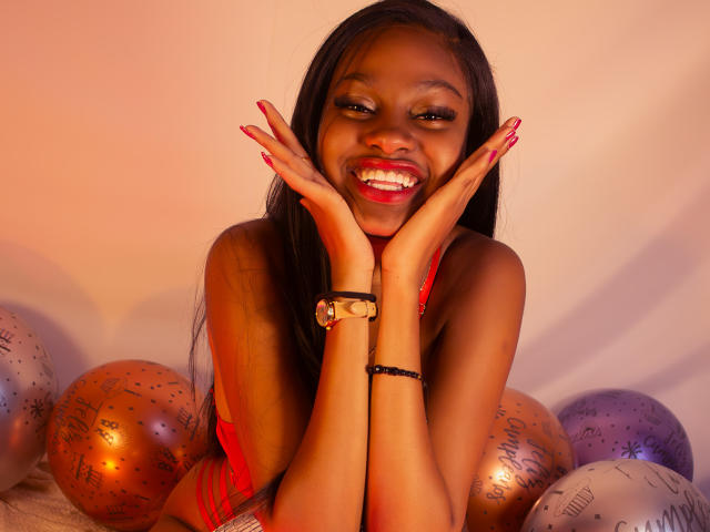 StormiHills with dark skin, long black hair, red lipstick, smiling, hands on cheeks, red nails, wearing red lingerie, gold and black bracelets, surrounded by orange and purple glitter balloons.