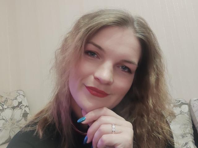 Lovelilii with wavy blonde hair, blue nail polish, red lipstick, smiling, black top, hand on chin, silver ring, pale skin, and blue eyes.