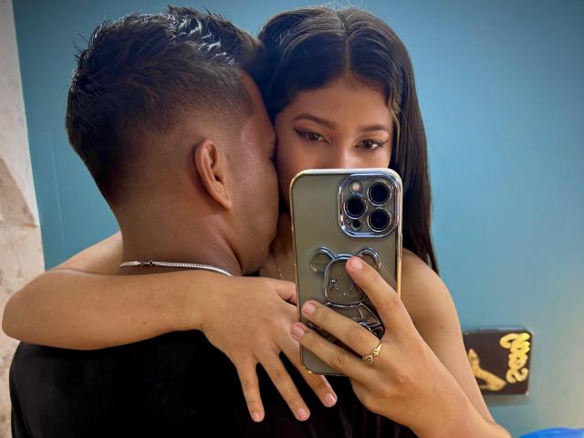 JonesAndJuly's face is near the camera, her brown eyes looking at it, while a man with short hair kisses her neck. She holds a phone with a bear case.