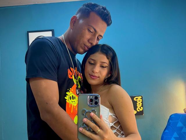 JonesAndJuly with tan skin and dark hair, in a white lace dress, leaning on a muscular tan-skinned guy in a black tee, taking a mirror selfie.