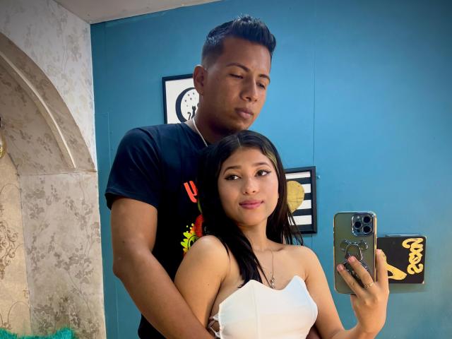 JonesAndJuly with short black hair stands behind a girl with long black hair, white strapless top, holding a phone. He's wearing a black t-shirt, arms around her.