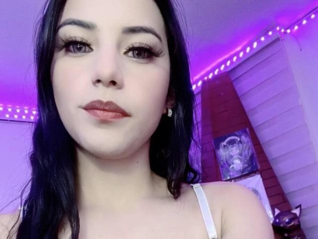 Arenne with pale skin, black hair, and red lipstick, wearing a white top, has long eyelashes and a slight smile, in a purple-lit room. Her tits are not visible.