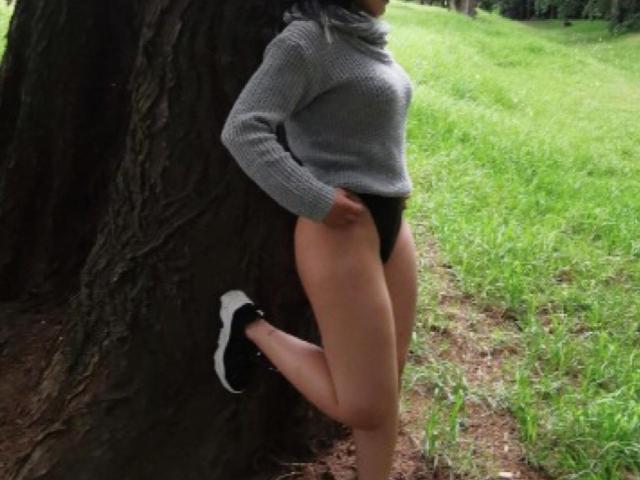 Arenne stands against a tree, wearing a grey sweater, black panties, and black-and-white sneakers, with her hand in her pocket, legs bent, and grassy field in background.