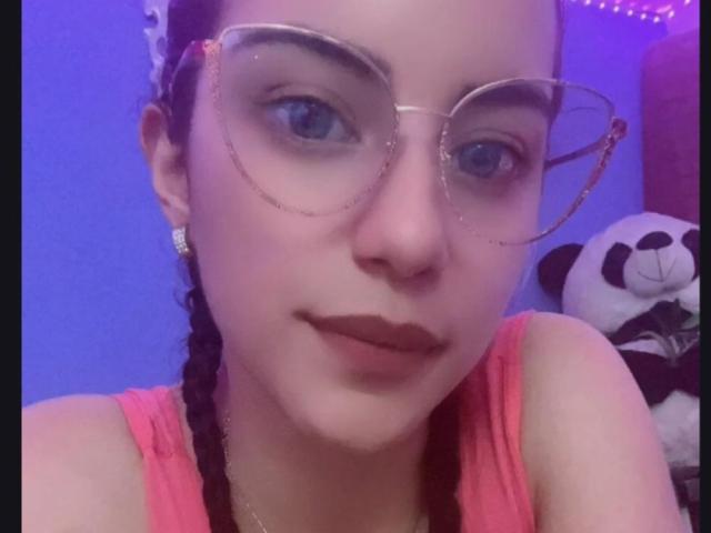 Arenne with glasses, pink tank top, dark braided hair, purple lighting, and a cute smile, showing her face and small tits, with a stuffed panda on the right.
