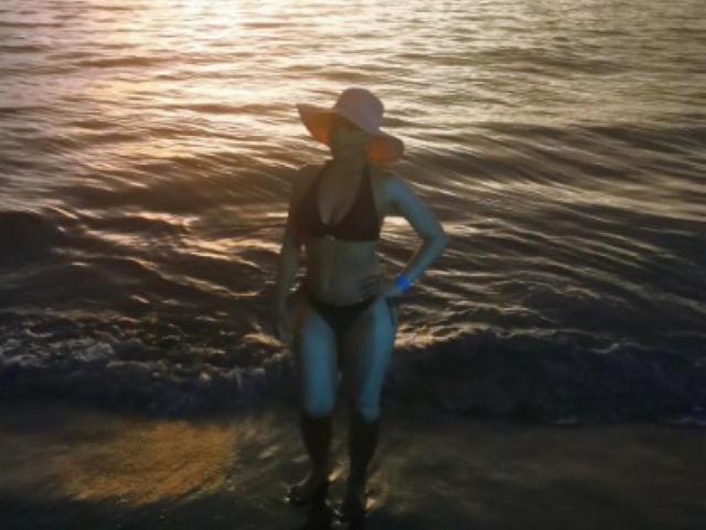 Arenne in a black bikini, wearing a sunhat and knee-high socks, standing at the water's edge with sunlight reflecting off the water. Her tits are small, ass is visible.