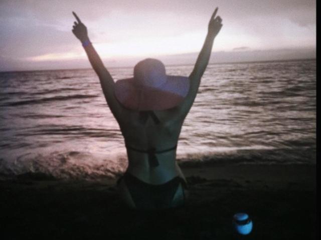 Arenne's back is to the camera, arms raised, wearing a black bikini with a sunhat, standing on a beach at sunset, holding a glowing bottle in her right hand.