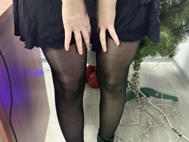 Laliinas schwarzer Rock, schwarze, durchsichtige Strumpfhosen und ihre Hände, die auf ihren Oberschenkeln ruhen. Ihre Beine sind leicht gespreizt, wodurch ihre mit Strumpfhosen bedeckte Vulva sichtbar ist. Sie steht vor einer Holzoberfläche..