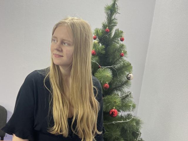 Laliina with long blonde hair, black top, looking to the side, standing next to a decorated Christmas tree with red and silver ornaments.