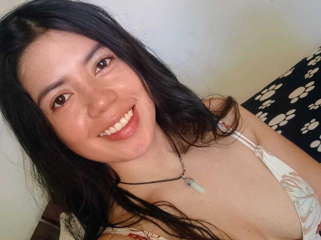 CristalDuport has long black hair, brown eyes, and a cute smile with her pink lips showing. She's wearing a white floral bra and a black necklace with a green pendant.