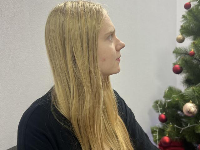 I capelli biondi e lisci di Laliina le arrivano oltre le spalle, indossa una maglietta nera e ha un piccolo segno rosso sulla guancia destra, ed è in piedi accanto a un albero di Natale decorato..