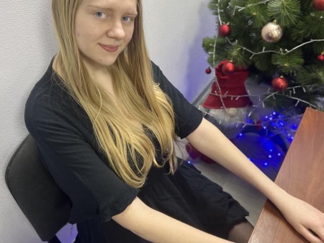 Laliina, con cabello rubio largo, vestido negro, sentada, sonriendo, con las manos sobre una mesa de madera, un árbol de Navidad con adornos y luces azules en el fondo..
