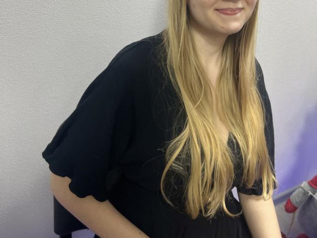 Laliina tem cabelos loiros longos, uma blusa preta e um sorriso discreto. Seus seios estão cobertos, e ela está em pé com as mãos na parte de trás das pernas. Ela é fofa..