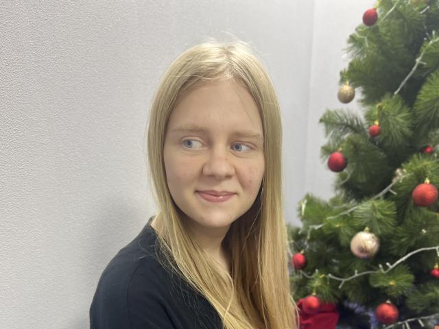 Laliina, con lunghi capelli biondi, occhi azzurri e un leggero sorriso, indossa una maglietta nera e si trova accanto a un albero di Natale decorato..
