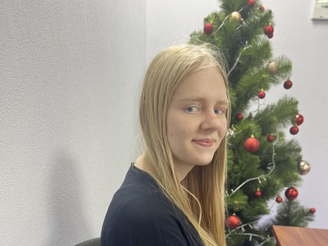 Laliina, con cabello rubio largo, ojos azules y una camisa negra, sonriendo a la cámara, está de pie junto a un árbol de Navidad decorado con adornos rojos y dorados..
