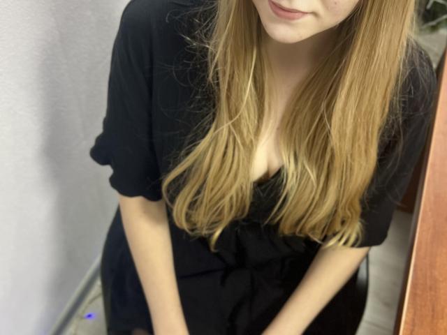 Laliina, com cabelos loiros longos, vestido preto e decote visível, sorrindo levemente, em pé com as mãos no vestido preto, em um quarto com uma porta de madeira..