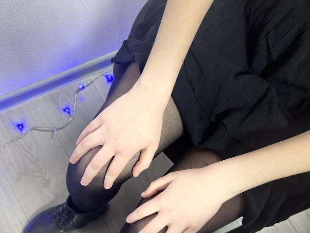 Laliina's pale hands rest on her black pantyhose-covered thighs, wearing a black dress, with blue fairy lights on the wooden floor, and black boots visible.