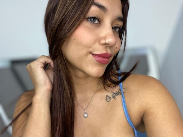 SophieDenisee tem cabelo castanho, um piercing no nariz, olhos azuis, batom rosa, uma tatuagem no ombro direito com a inscrição "E. Pine" e um colar de prata com um coração..