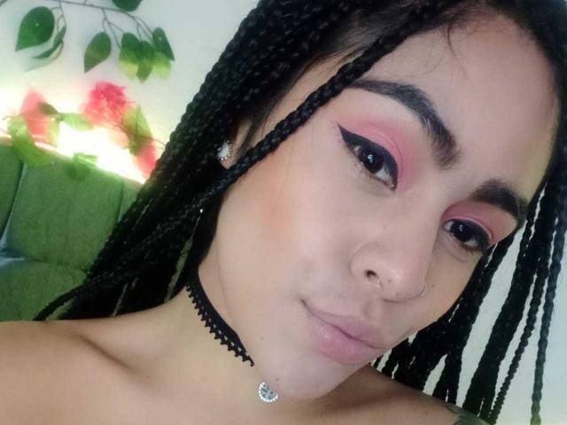 StellaBeckker with braided hair, pink eyeshadow, black choker, nose ring, and earrings, showing a subtle smile, with visible tits in the foreground.