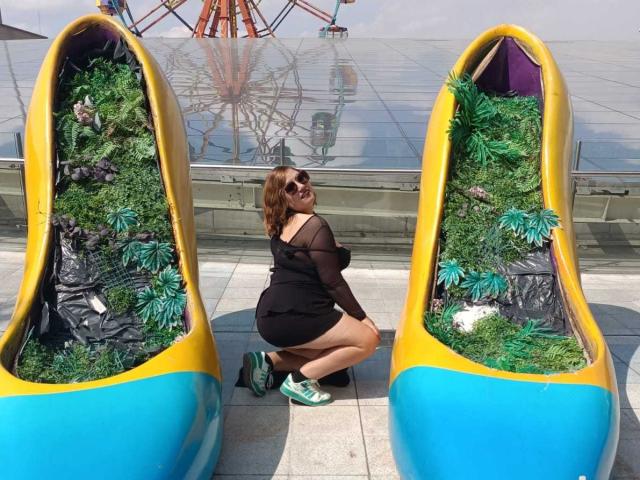 NatashaChaus, with brown hair and sunglasses, kneels in front of two yellow and blue planters with green foliage. She's wearing a black sheer dress and green sneakers.