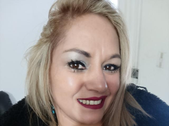 NatashaChaus has blonde hair, blue eyeshadow, thick black eyelashes, red lipstick, and is smiling with her teeth showing. She's wearing a black fur-like garment.