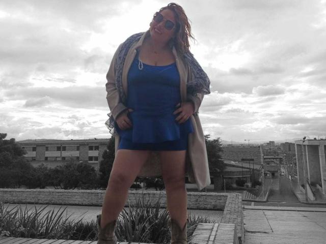 NatashaChaus stands with hands on hips, wearing a blue dress, tan coat, and sunglasses, her ass slightly exposed, and boots on a rooftop. Her red hair is wet.