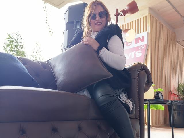 NatashaChaus, with red hair and sunglasses, sits on a brown tufted sofa, hugging a black pillow, wearing a white long-sleeve shirt and black leather pants.