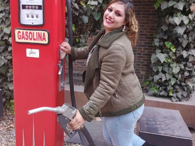 NatashaChaus with curly brown hair, green jacket, and blue jeans, smiling while pumping gas from a red gas pump, holding the nozzle with her right hand.