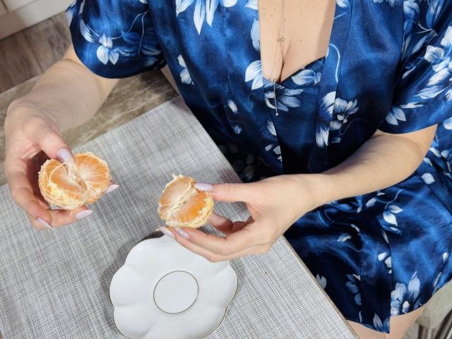 LisaTreds, met bleke huid, draagt een blauwe jurk met bloemenprint en houdt in elke hand een halve grapefruit vast. Haar vingers hebben bleekroze nagels. Onder haar bevindt zich een wit bord met een cirkelvormig ontwerp..