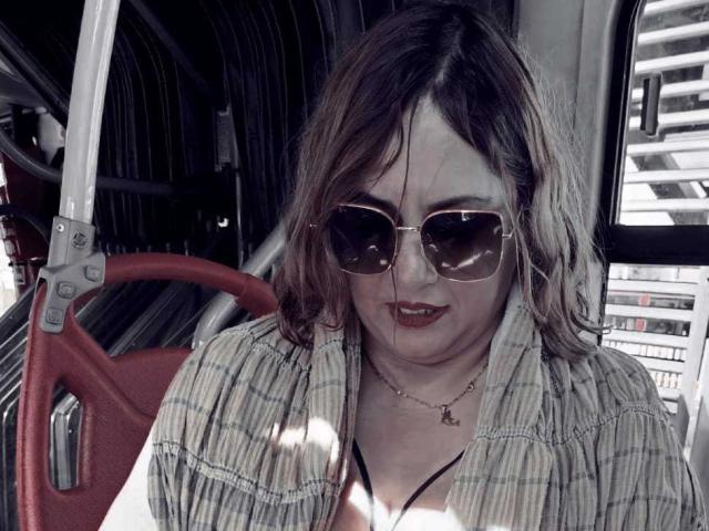 NatashaChaus with messy brown hair, wearing sunglasses, a plaid shirt, and a black necklace, sitting in a bus, smiling slightly, showing her small tits.