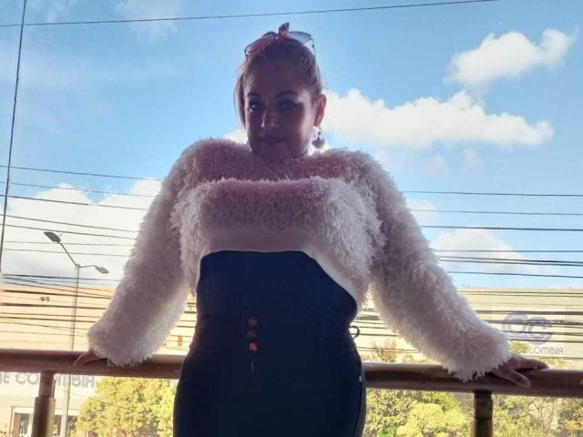 NatashaChaus, with pink fluffy sweater, black high-waisted pants, and button details, leaning on a railing, blue sky and clouds in background, sunglasses on head, standing confidently.