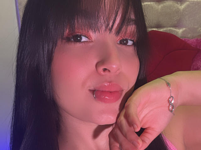 SarahGood with black hair, pink eyeshadow, and glossy pink lips, biting her lower lip. Her right hand, with a silver bracelet, rests near her face.