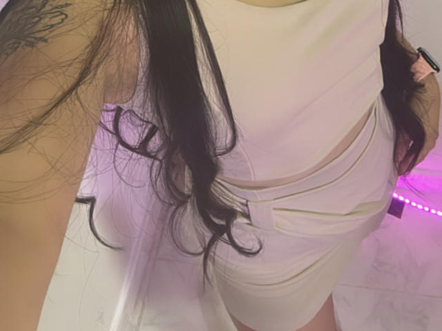 SarahGood indossa un abito bianco aderente con aperture, ha i capelli lunghi e neri, un tatuaggio sul braccio destro e una luce rosa sullo sfondo. Il suo sedere è leggermente visibile attraverso l'abito..