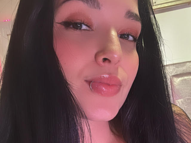 SarahGood's face with black hair, pinkish-red lipstick, and glossy pout, sticking out her tongue slightly. She has bold eyeliner and a light pink blush on her cheeks.