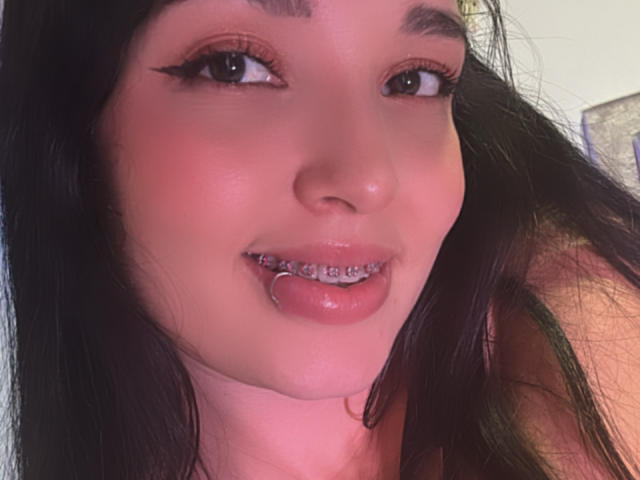 SarahGood has black hair, brown eyes, and is sticking her tongue out with metal braces. She has pink lips and winged eyeliner. Her skin is fair.