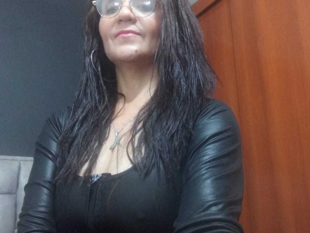 SamantaaFox with wet black hair, glasses, and black leather jacket, showing her tits in a tight top, standing with a slight smile, wooden background.
