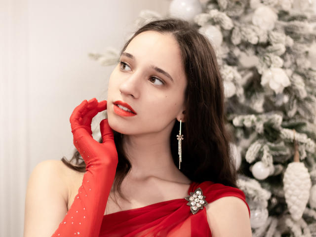 StacieLove with red lipstick, wearing red glitter gloves and a red sheer dress, looking to the side, has a cross earring, and a snow-covered Christmas tree in the background.