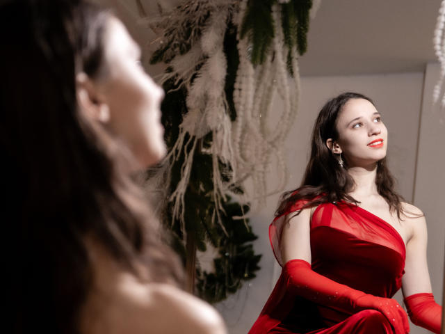 StacieLove vestindo um vestido vermelho com luvas combinando, olhando para um espelho com o traseiro levemente virado, cabelo escuro, batom vermelho e um sorriso confiante. Reflexo no espelho..
