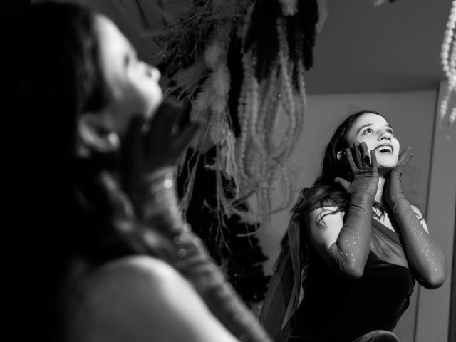 Black-and-white photo of StacieLove with long hair, laughing, wearing sparkly gloves and a dress, hands near face, blurred profile of another StacieLove in foreground.