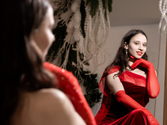 StacieLove com um vestido vermelho e luvas, sentada em frente a um espelho, olhando para o reflexo, com a mão no queixo, batom vermelho, cabelo longo e escuro, elegante, sexy, brincos de pérola, iluminação suave..