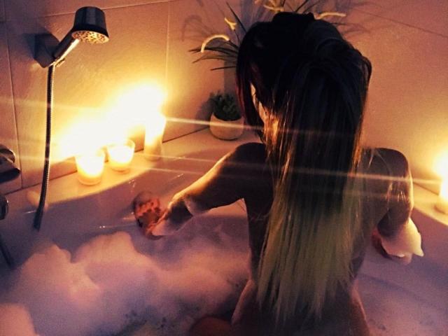 LillaGrey, con su largo cabello rubio, está sentada en una bañera llena de burbujas, alcanzando su pequeño trasero, rodeada de velas y un cabezal de ducha. Iluminación cálida y tenue..