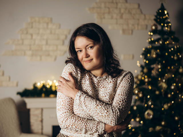 LolaHikls with medium tits, wearing a white lace sweater, arms crossed, smiling, brown wavy hair, standing near a decorated Christmas tree, warm indoor lighting.