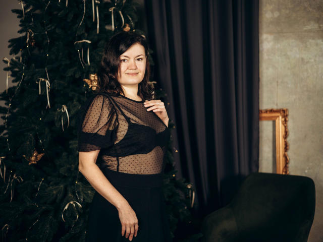 LolaHikls, con i capelli scuri e ondulati, sorride, indossando una maglietta nera trasparente che rivela i suoi piccoli seni, in piedi accanto a un albero di Natale in una stanza poco illuminata..
